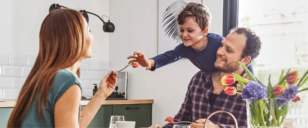 Familie sitzt gemeinsam am Holztisch in moderner Küche. Eltern und Kind genießen eine Mahlzeit in heller Wohnküche mit natürlichem Licht, Blumen und wohnlicher Atmosphäre. Küche als Mittelpunkt des Familienlebens.
